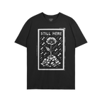 The Still Here T-shirt features a linocut-style poppy growing through rubble, surrounded by falling shapes, symbolizing resil