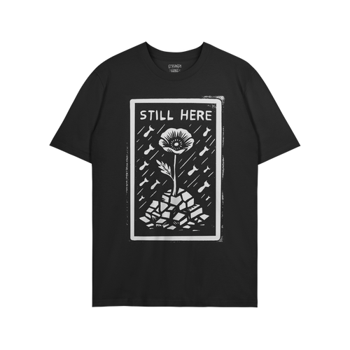 The Still Here T-shirt features a linocut-style poppy growing through rubble, surrounded by falling shapes, symbolizing resil