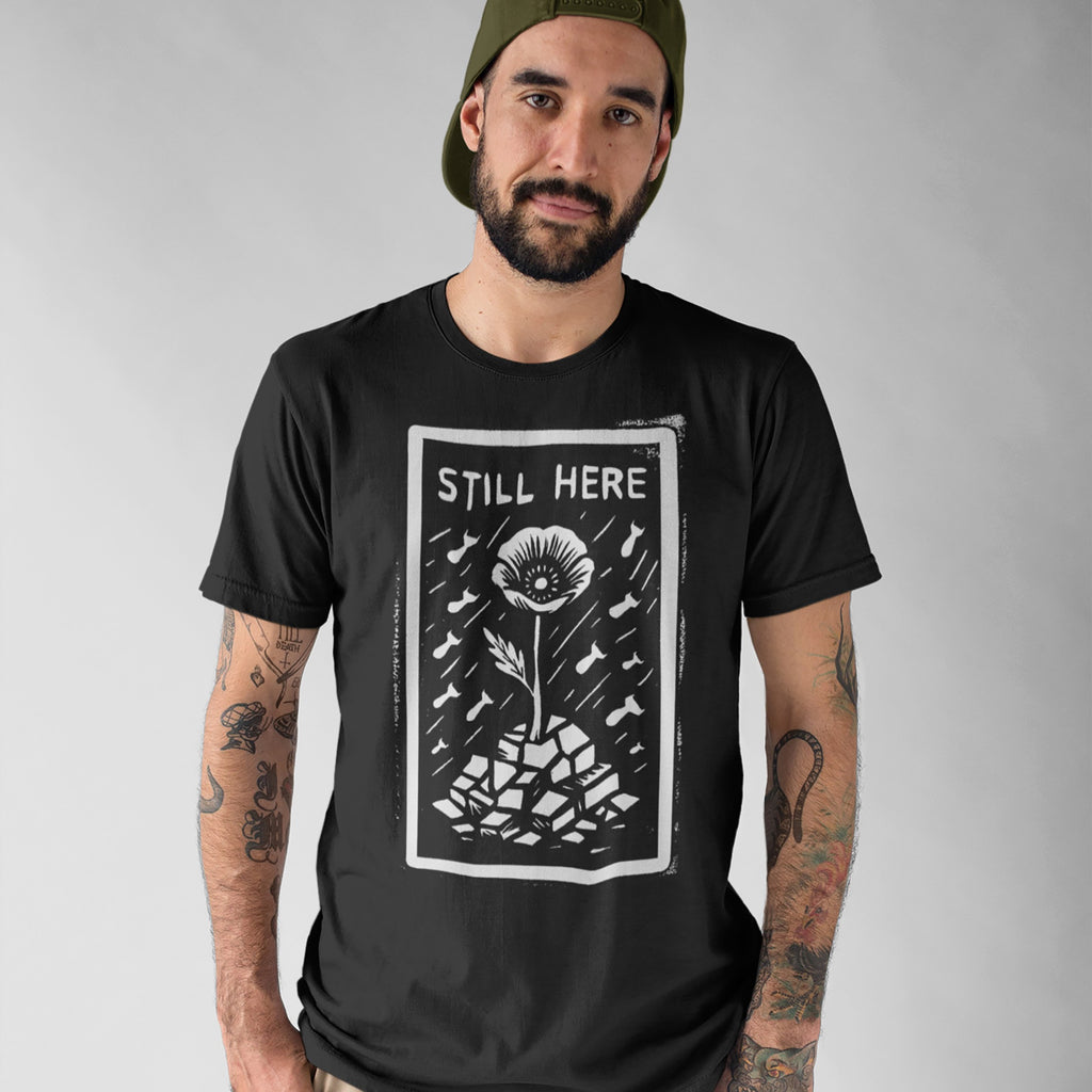 A bearded man models the Still Here T-shirt, featuring a woodcut-style graphic of a resilient flower amidst falling bombs, symbolizing resistance against oppression. He's against a neutral gray backdrop.