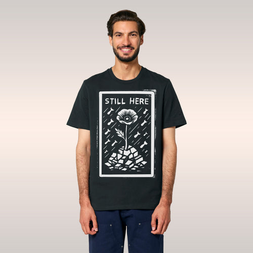 A bearded man models the Still Here T-shirt, featuring a woodcut-style graphic of a resilient flower amidst falling bombs, sy