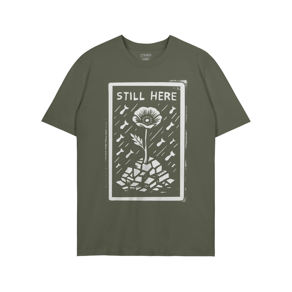 Still Here T-shirt features a bold graphic of a flower rising through rubble with bombs falling, symbolizing resilience and p