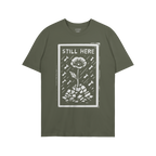 Still Here T-shirt features a bold graphic of a flower rising through rubble with bombs falling, symbolizing resilience and p
