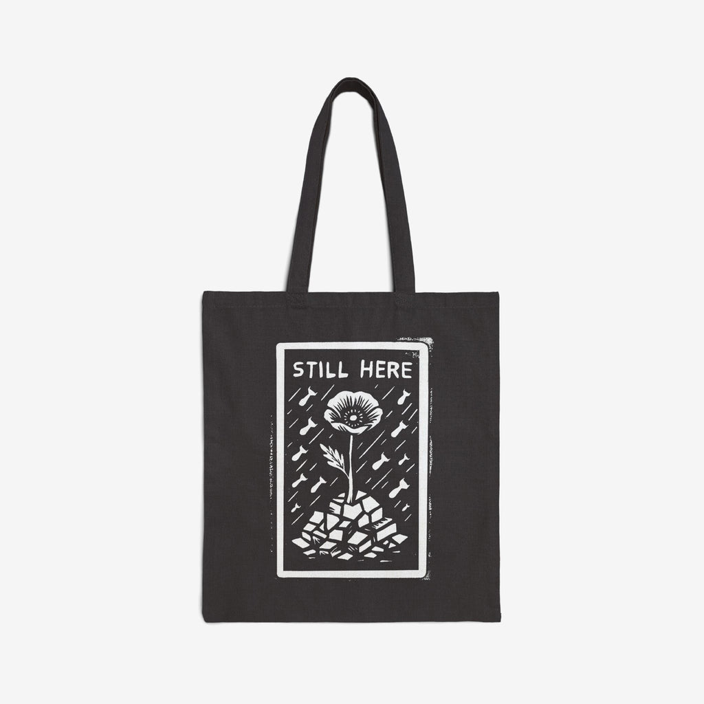 The Still Here Tote Bag displays a white hand-drawn flower rising from stones amid falling rain, symbolizing resilience against adversity. Ideal for activists, it's a powerful statement against oppression.