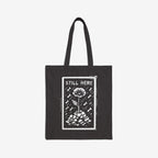 The Still Here Tote Bag displays a white hand-drawn flower rising from stones amid falling rain, symbolizing resilience against adversity. Ideal for activists, it's a powerful statement against oppression.