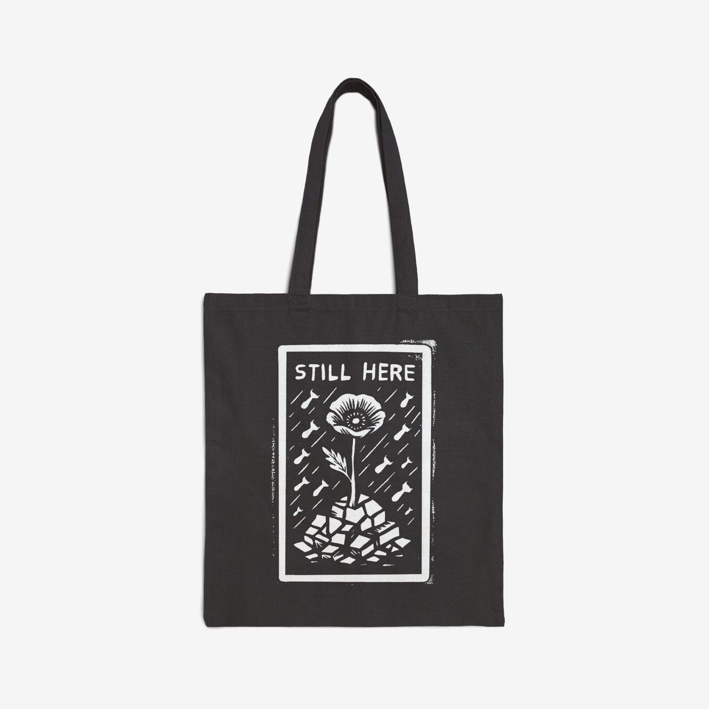 The Still Here Tote Bag displays a white hand-drawn flower rising from stones amid falling rain, symbolizing resilience against adversity. Ideal for activists, it's a powerful statement against oppression.