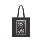The Still Here Tote Bag displays a white hand-drawn flower rising from stones amid falling rain, symbolizing resilience again