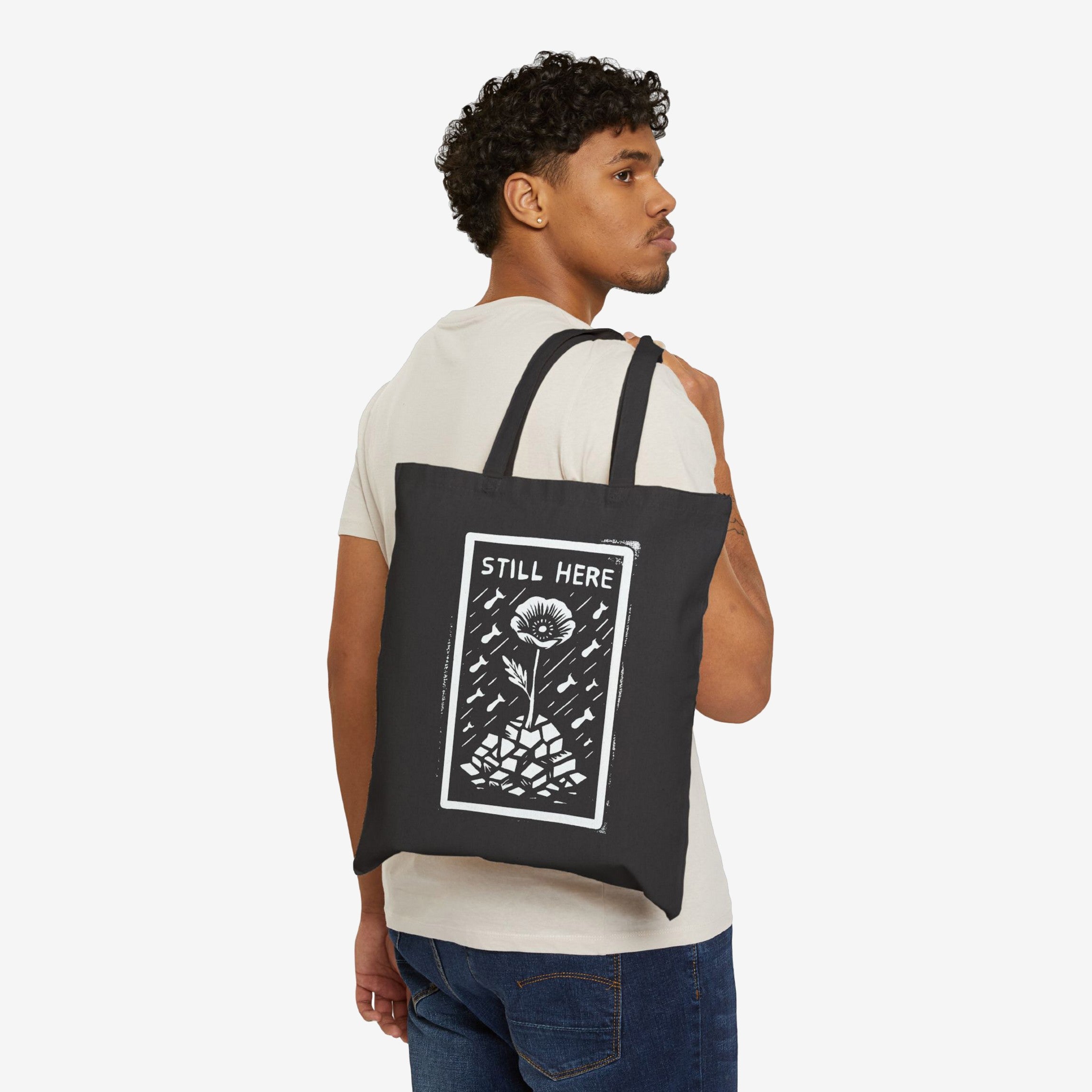 Still Here Tote Bag: A young man carries a black canvas tote with a flower emerging from rubble design, symbolizing resilience. Features block letters STILL HERE. Ideal for supporters of human rights and equality.
