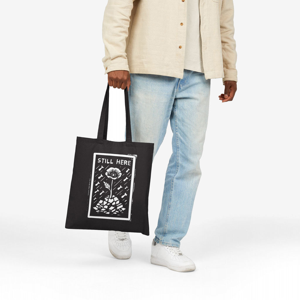 Still Here Tote Bag with a flower graphic symbolizing resilience amid adversity, held by a person in jeans and white sneakers, showcasing a message of enduring strength and solidarity.