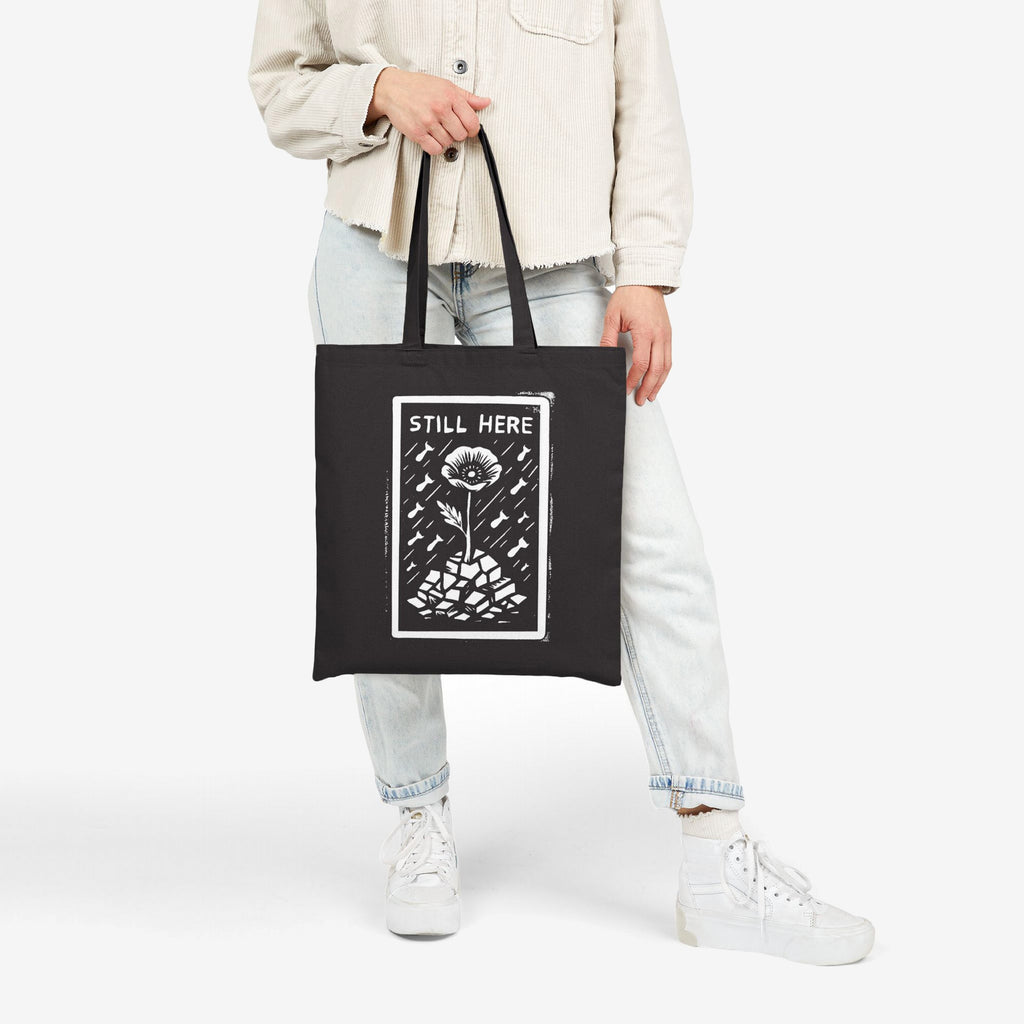 Still Here Tote Bag featuring a flower in cracked earth under rain, symbolizing resilience through adversity, held by a person wearing light-wash jeans and white sneakers. A statement piece for activism.