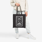Still Here Tote Bag featuring a flower in cracked earth under rain, symbolizing resilience through adversity, held by a person wearing light-wash jeans and white sneakers. A statement piece for activism.