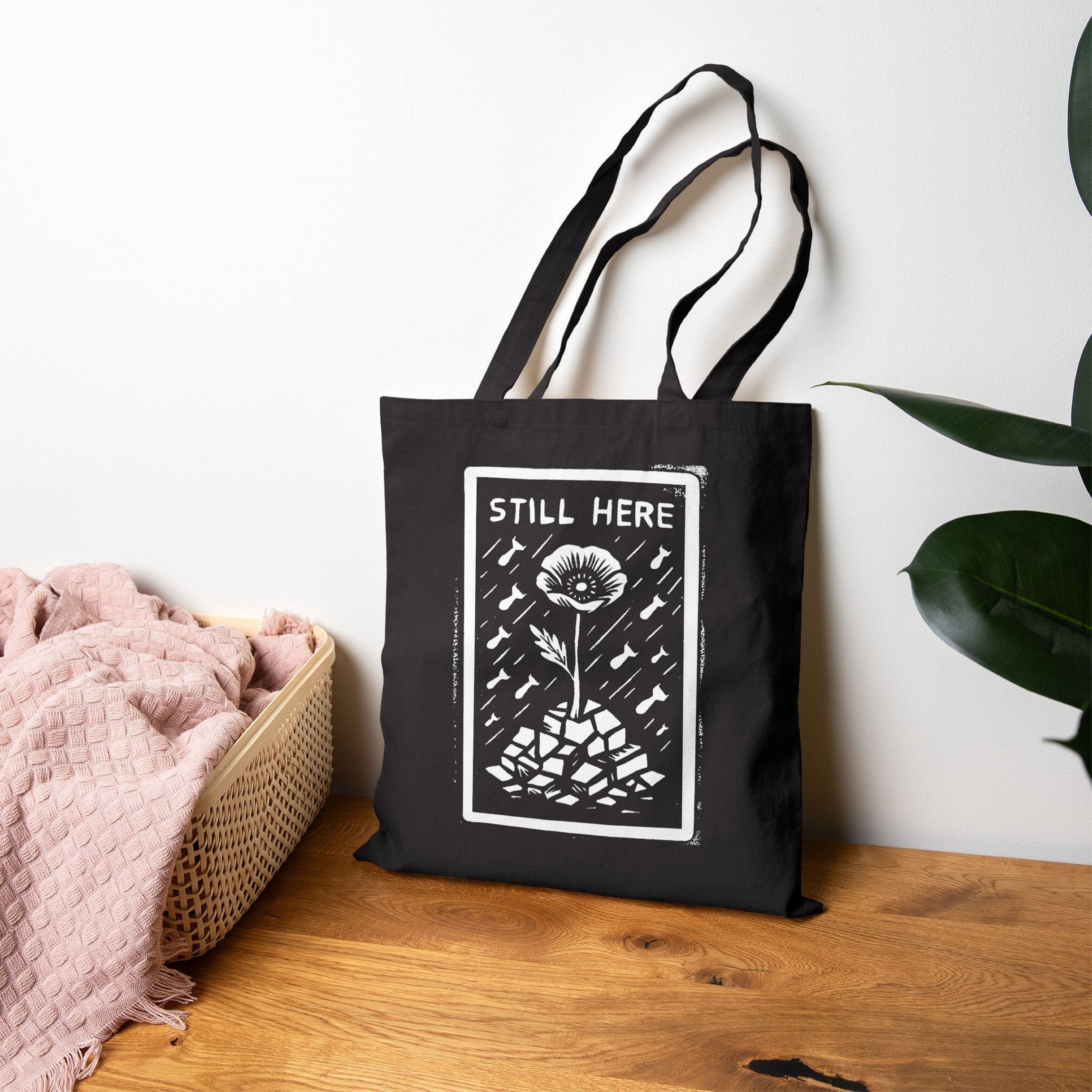 The Still Here Tote Bag features a linocut-style graphic of a flower amidst rubble, symbolizing resilience. Positioned against a wall, it embodies strength amid adversity, perfect for advocates of justice and equality.
