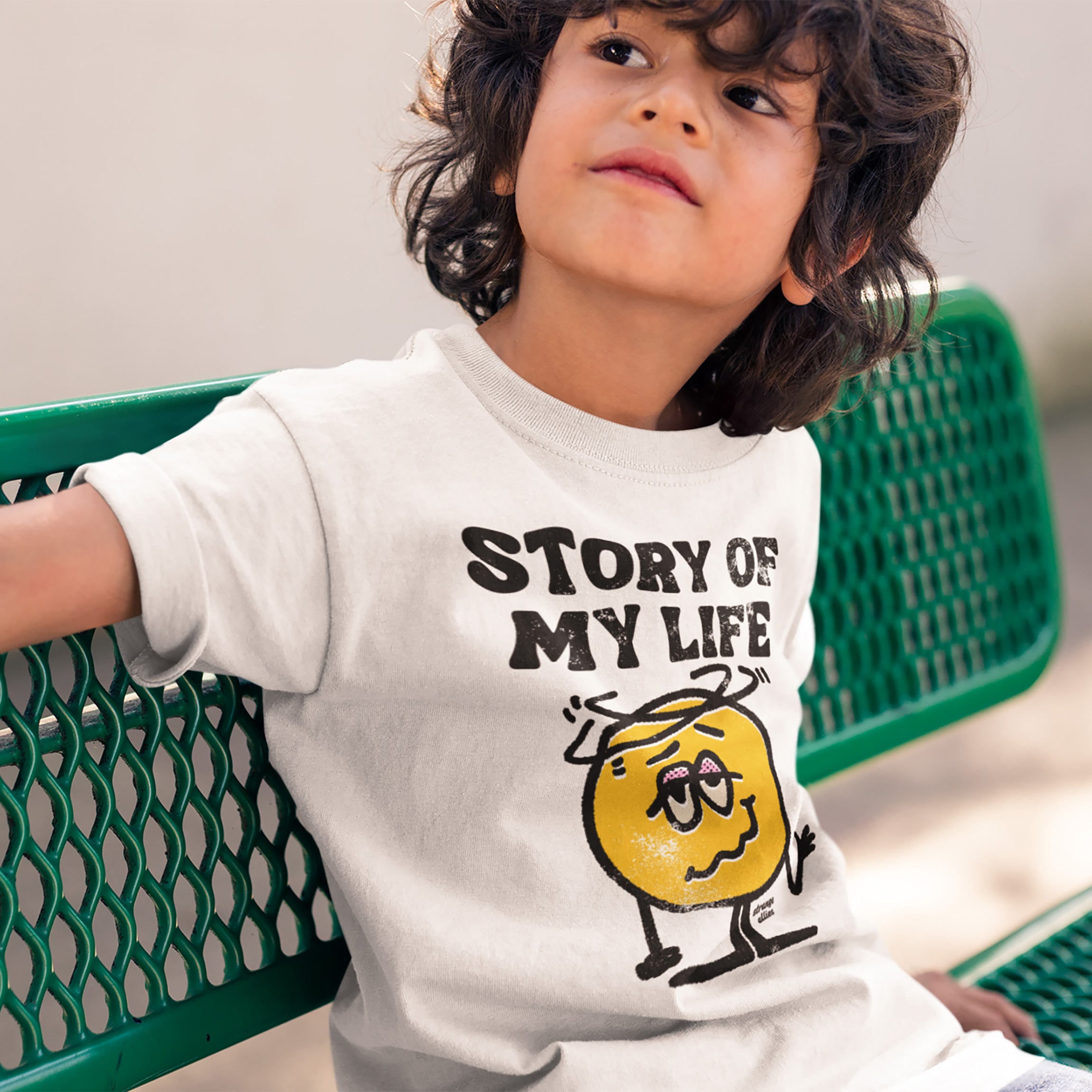 Child wearing the Story Of My Life T-shirt featuring a sleepy-eyed cartoon, sitting on a bench outdoors, embodying a relatable, humorous take on daily chaos.