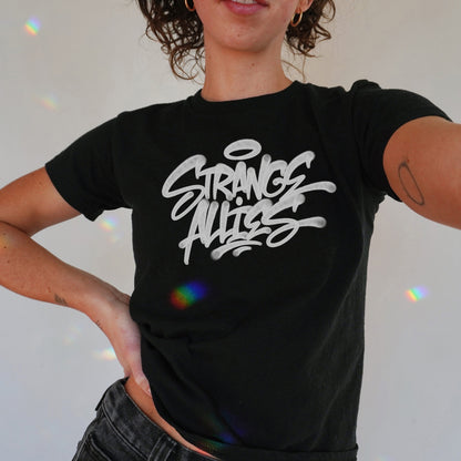 Person wearing the Strange Allies Handstyle Women's Baby Tee, featuring bold graffiti-style text and a halo motif, embodying an activist spirit. The tee is cropped, fitted, and paired with minimal tattoos and gold hoops.
