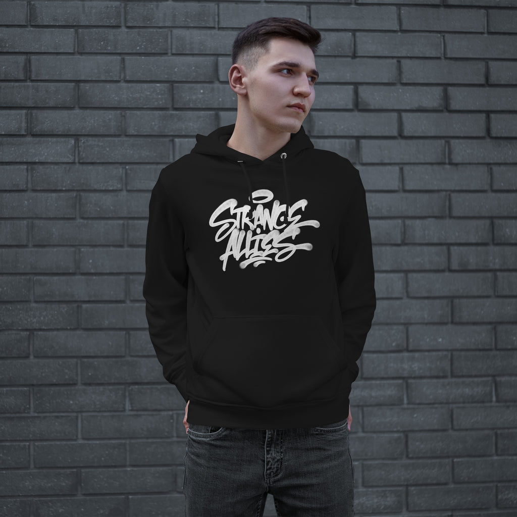 Young man wearing the Strange Allies Handstyle Hoodie, featuring a bold graffiti-style logo STRANGE ALLIES across the chest, standing casually with hands in pockets against a gray brick wall.