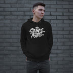 Young man wearing the Strange Allies Handstyle Hoodie, featuring a bold graffiti-style logo STRANGE ALLIES across the chest, standing casually with hands in pockets against a gray brick wall.
