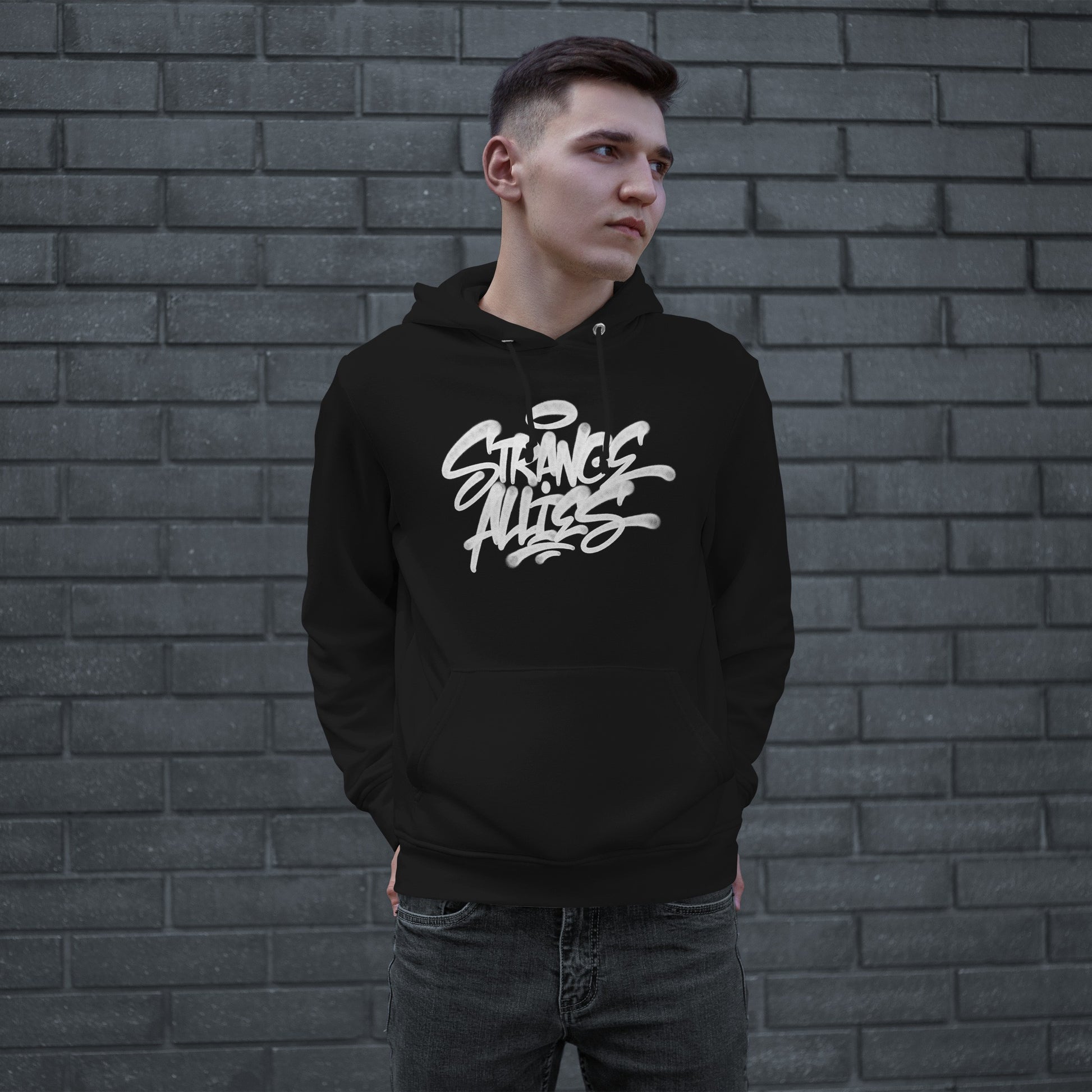 Young man wearing the Strange Allies Handstyle Hoodie, featuring a bold graffiti-style logo STRANGE ALLIES across the chest, standing casually with hands in pockets against a gray brick wall.