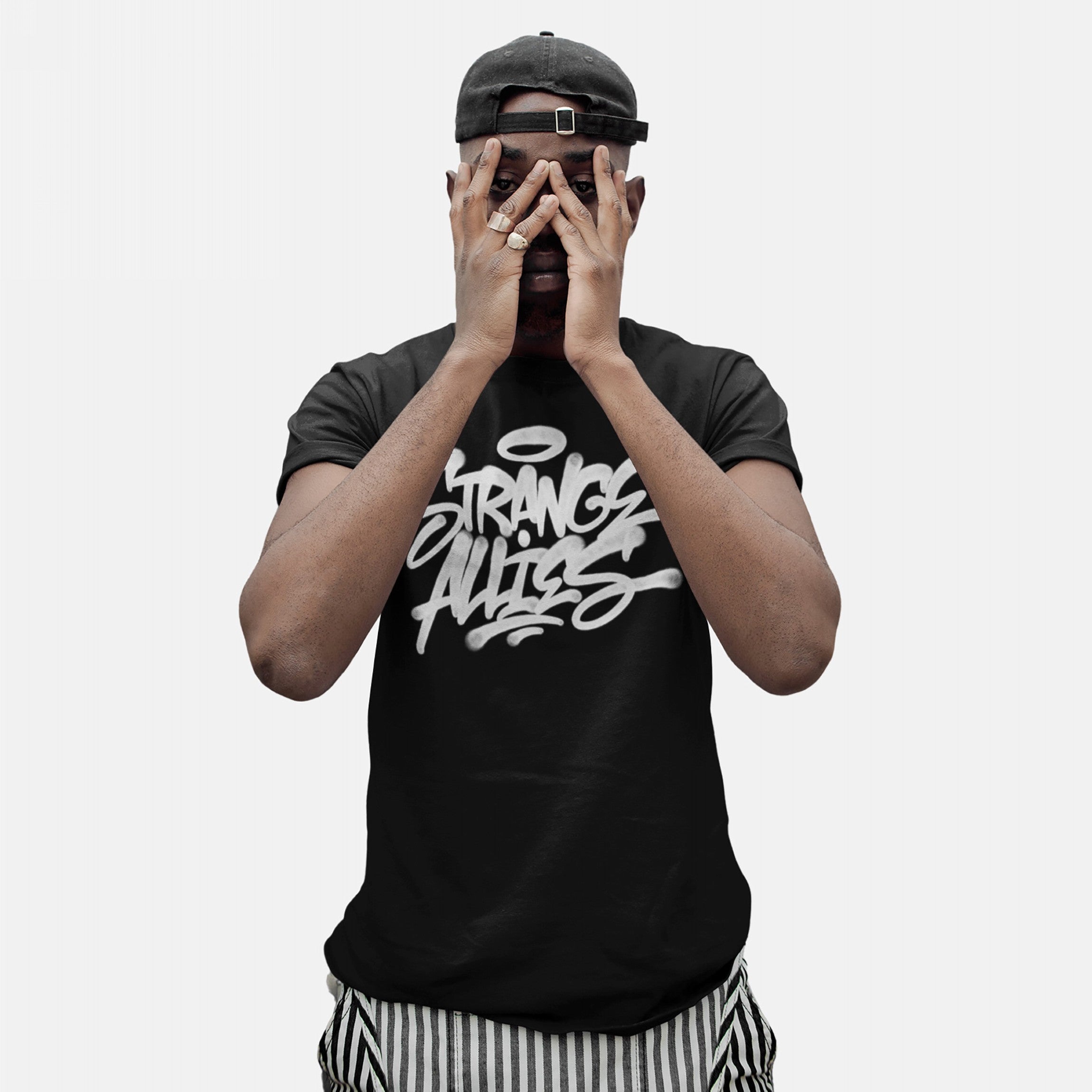 Young man in a black Strange Allies Handstyle T-shirt with graffiti-style lettering, wearing a backwards baseball cap, holding hands over his face, paired with black-and-white striped trousers.