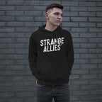 Young man wearing the Strange Allies Hoodie, featuring bold white STRANGE ALLIES™ text, stands confidently with hands in pockets, embodying the brand's essence of unity and defiance against a gray brick wall.