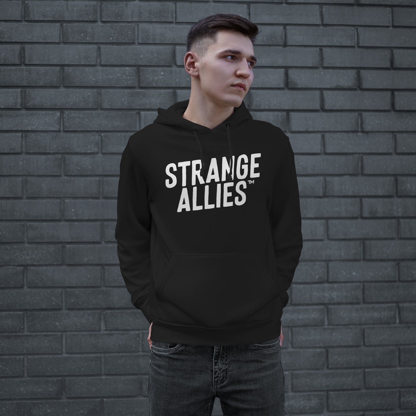 Young man wearing the Strange Allies Hoodie, featuring bold white STRANGE ALLIES™ text, stands confidently with hands in pockets, embodying the brand's essence of unity and defiance against a gray brick wall.