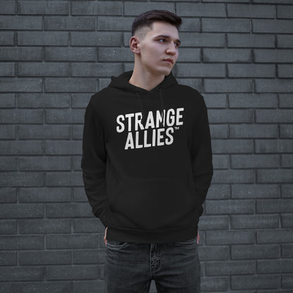 Young man wearing the Strange Allies Hoodie, featuring bold white STRANGE ALLIES™ text, stands confidently with hands in pockets, embodying the brand's essence of unity and defiance against a gray brick wall.