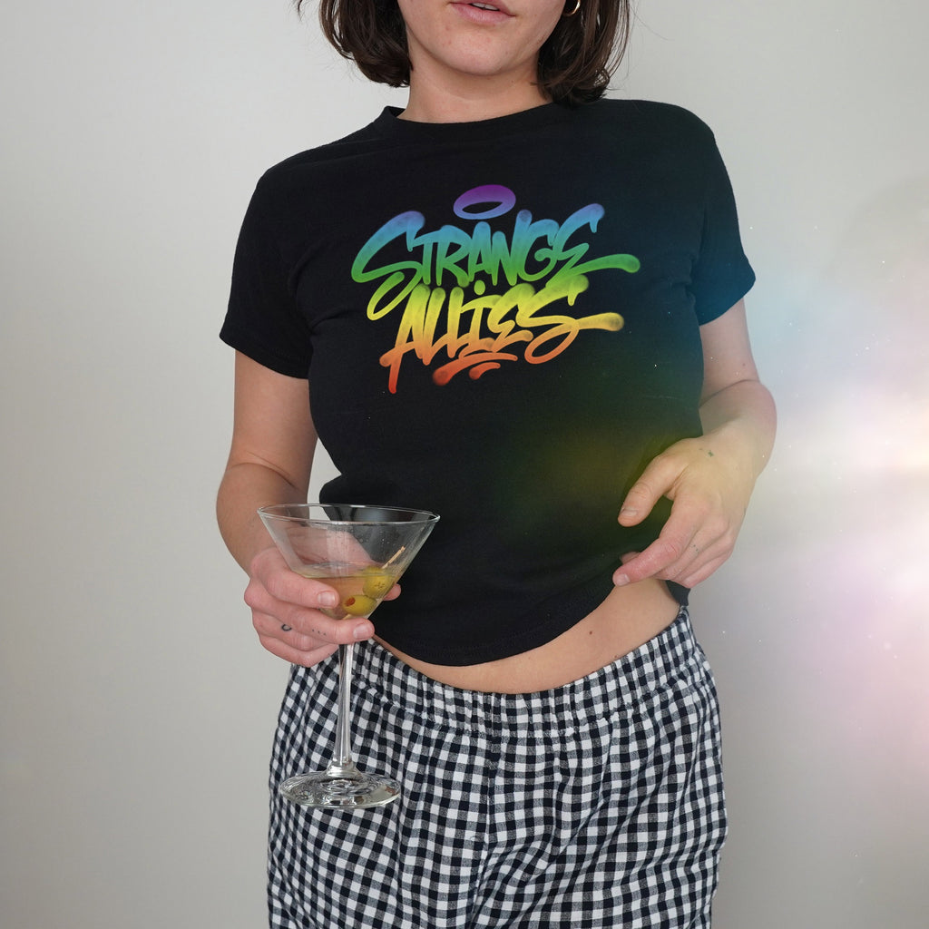 A person wearing the Strange Allies Rainbow Handstyle Women's Baby Tee, featuring rainbow-gradient lettering, paired with checkered pants, holds a martini glass, embodying vibrant allyship and LGBTQ+ pride.