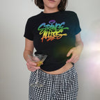 A person wearing the Strange Allies Rainbow Handstyle Women's Baby Tee, featuring rainbow-gradient lettering, paired with checkered pants, holds a martini glass, embodying vibrant allyship and LGBTQ+ pride.