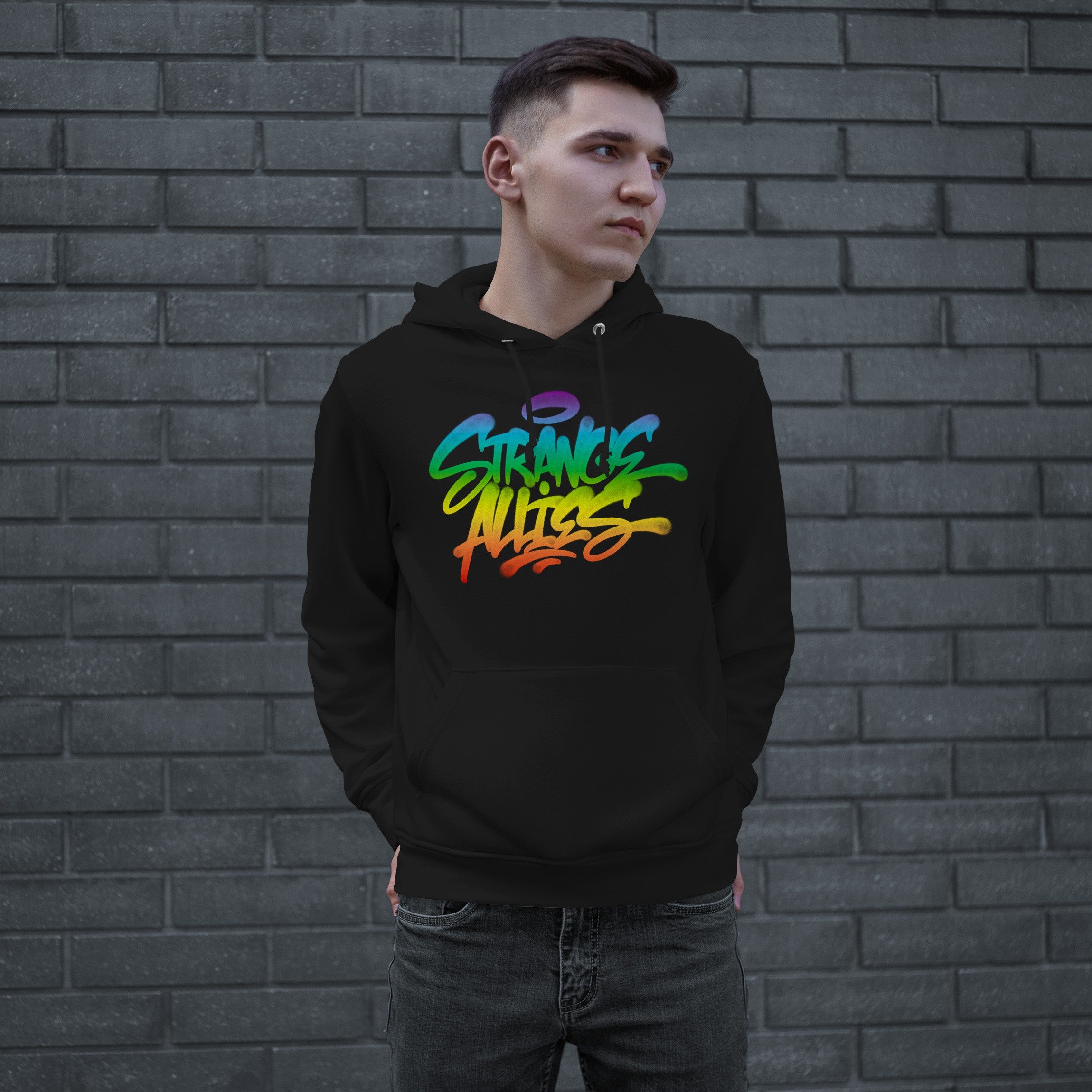 Young man wearing the Strange Allies Rainbow Handstyle Hoodie, featuring vibrant graffiti-style lettering. He stands against a dark grey brick wall, embodying an urban streetwear vibe with hands in the front pouch pocket.
