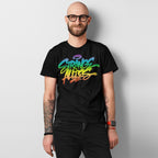 Smiling man wearing a Strange Allies Rainbow Handstyle T-shirt with vibrant graffiti script, showcasing tattoos and a red smartwatch, embodying bold individuality and inclusivity.