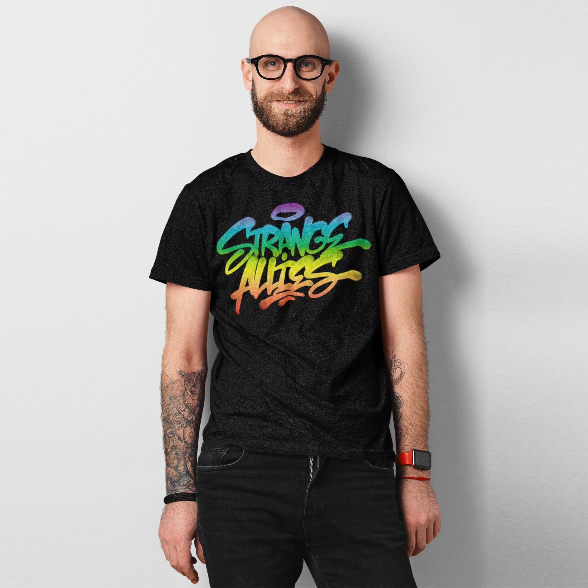 Smiling man wearing a Strange Allies Rainbow Handstyle T-shirt with vibrant graffiti script, showcasing tattoos and a red smartwatch, embodying bold individuality and inclusivity.