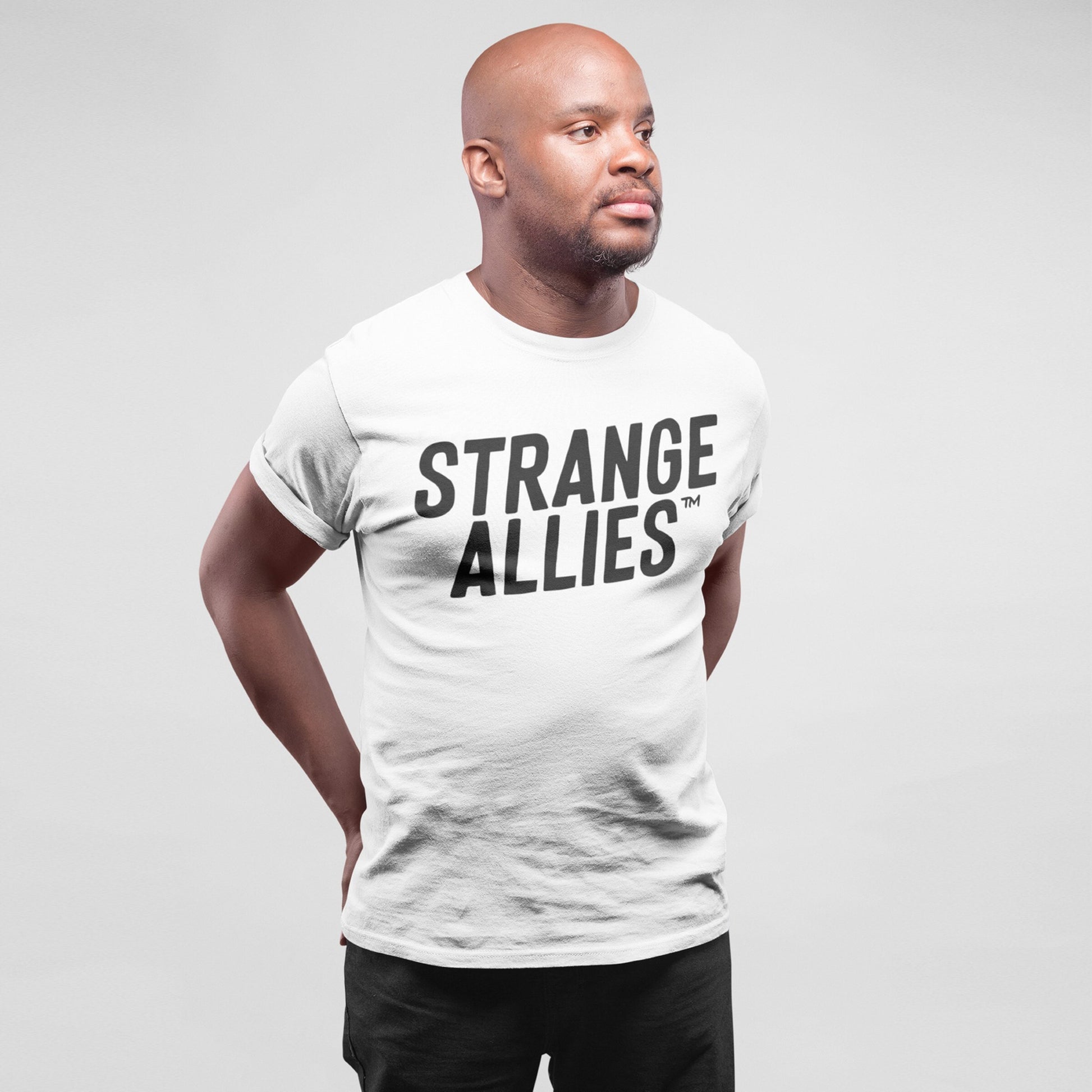 Bald man models the Strange Allies T-shirt, featuring bold text on a plain white tee, embodying a message of embracing uniqueness and individuality. Studio portrait highlights relaxed, confident style.
