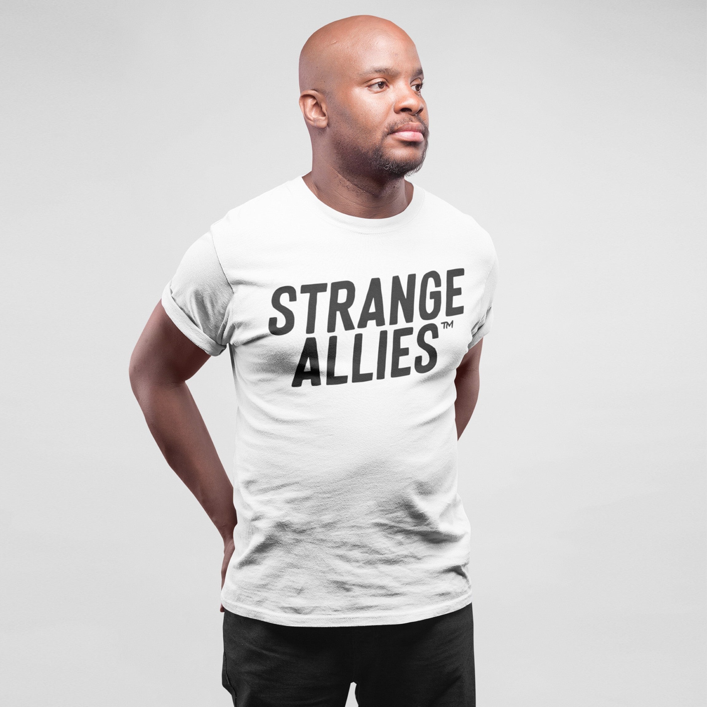 Bald man models the Strange Allies T-shirt, featuring bold text on a plain white tee, embodying a message of embracing uniqueness and individuality. Studio portrait highlights relaxed, confident style.