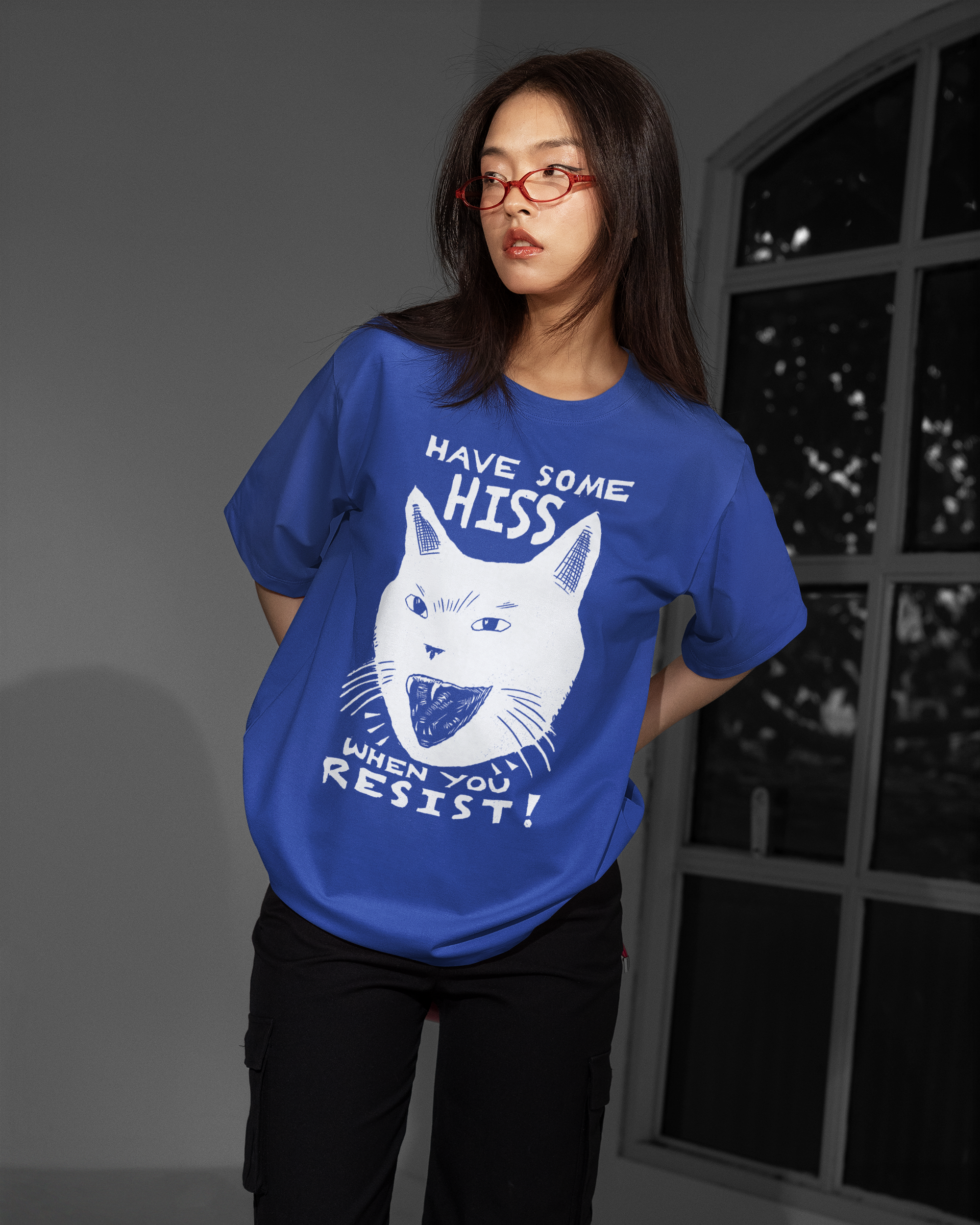 A woman modeling a blue graphic t-shirt with a white cat illustration and the text 'HAVE SOME HISS WHEN YOU RESIST!' in a moody, dark studio setting.