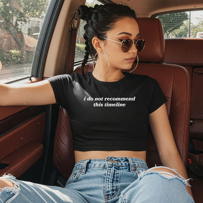 A young woman wears the Timeline Women's Baby Tee, featuring the phrase i do not recommend this timeline, paired with light-wash ripped jeans and aviator sunglasses, lounging in a car's leather interior.