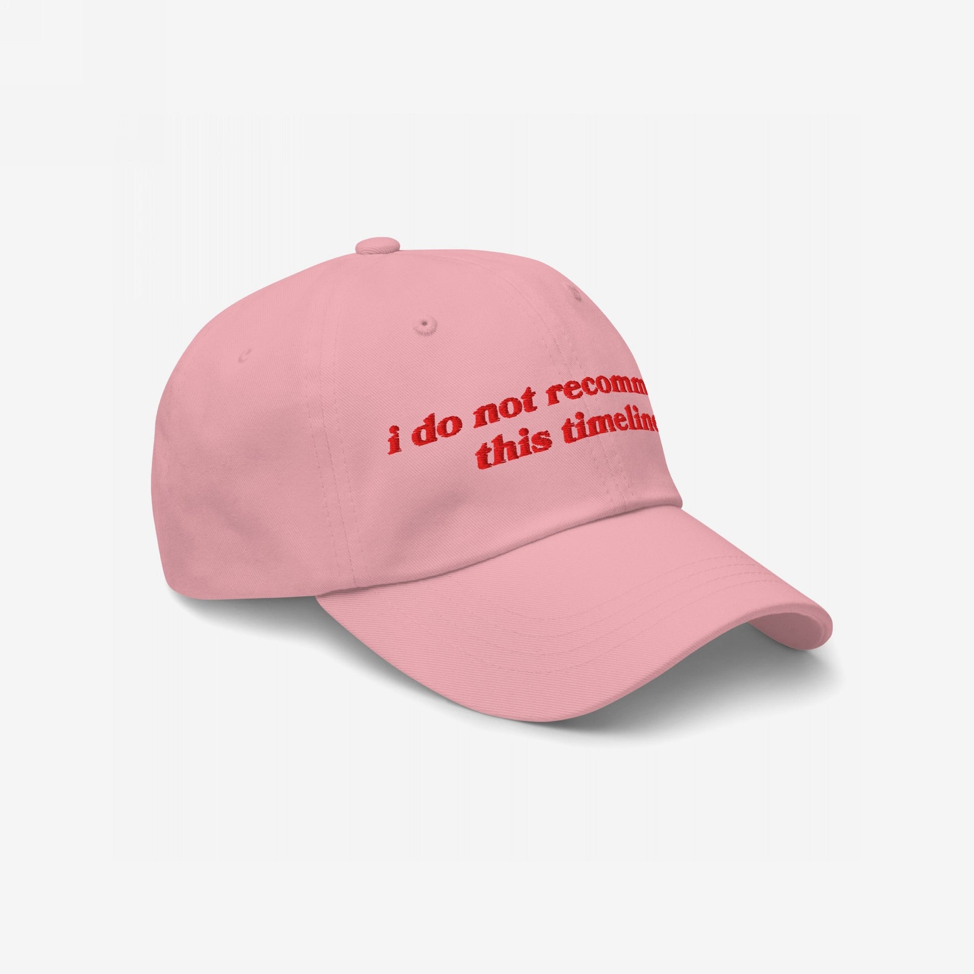 The Timeline Hat features bold red embroidery stating i do not recommend this timeline on a soft pink dad hat, capturing a sarcastic stance on current events with vintage-inspired lettering.