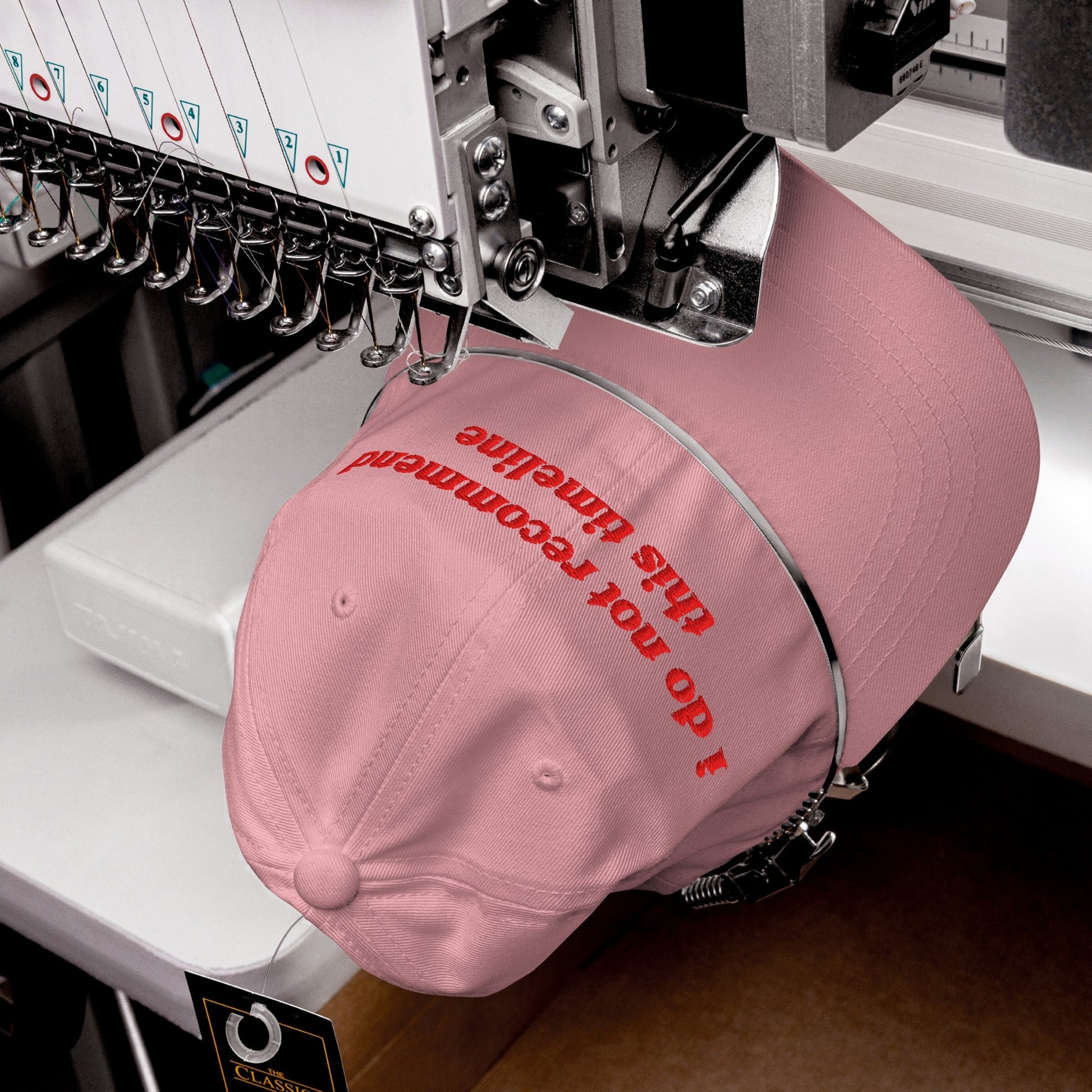 The Timeline Hat in production: An industrial embroidery machine stitches I do not recommend this timeline onto a pink cap, highlighting its sarcastic, politically conscious message.