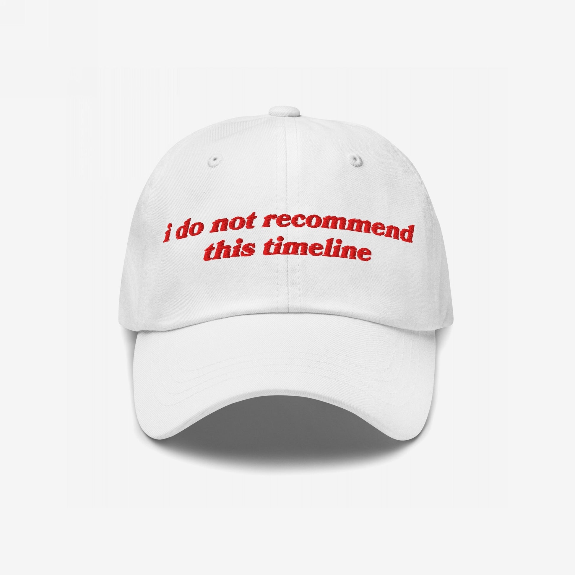 White baseball-style Timeline Hat with red embroidered text, I Do Not Recommend This Timeline, reflecting politically charged, sarcastic sentiments. Features a curved brim and ventilation eyelets, embodying a soft fit for hard truths.