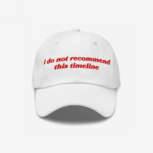 White baseball-style Timeline Hat with red embroidered text, I Do Not Recommend This Timeline, reflecting politically charged, sarcastic sentiments. Features a curved brim and ventilation eyelets, embodying a soft fit for hard truths.