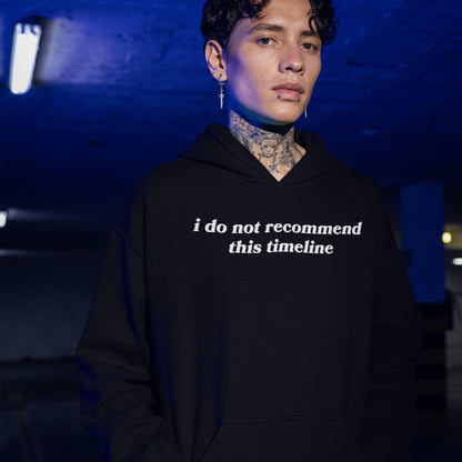 A young person in a dim parking garage wears a black Timeline Shirt hoodie, showcasing neck tattoos and piercings. The shirt humorously critiques modern life, aligning with a bold, expressive style.