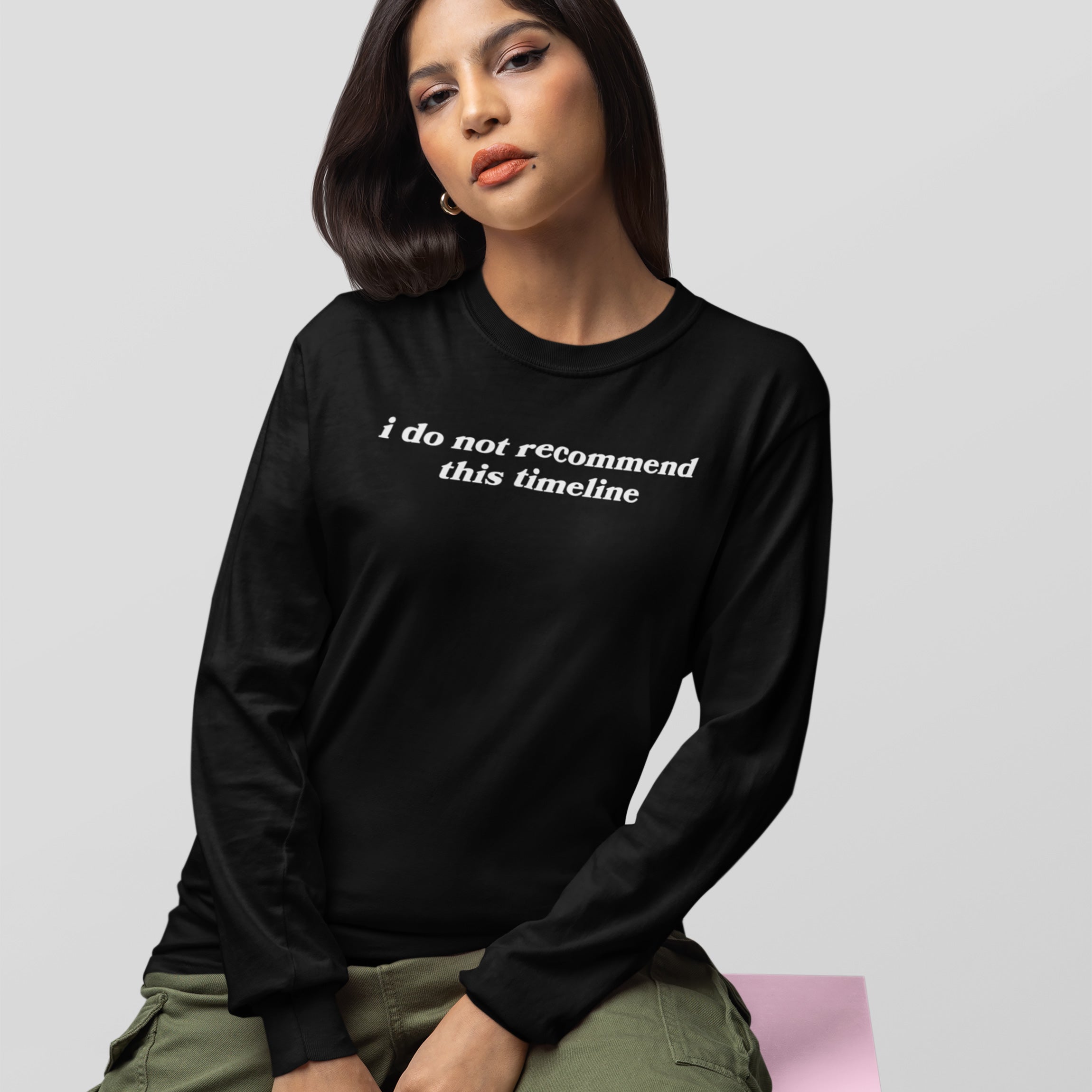 Young woman in black long-sleeve Timeline Shirt with white text, paired with olive cargo pants, seated on a pink block, embodying humor and critique for today's world.