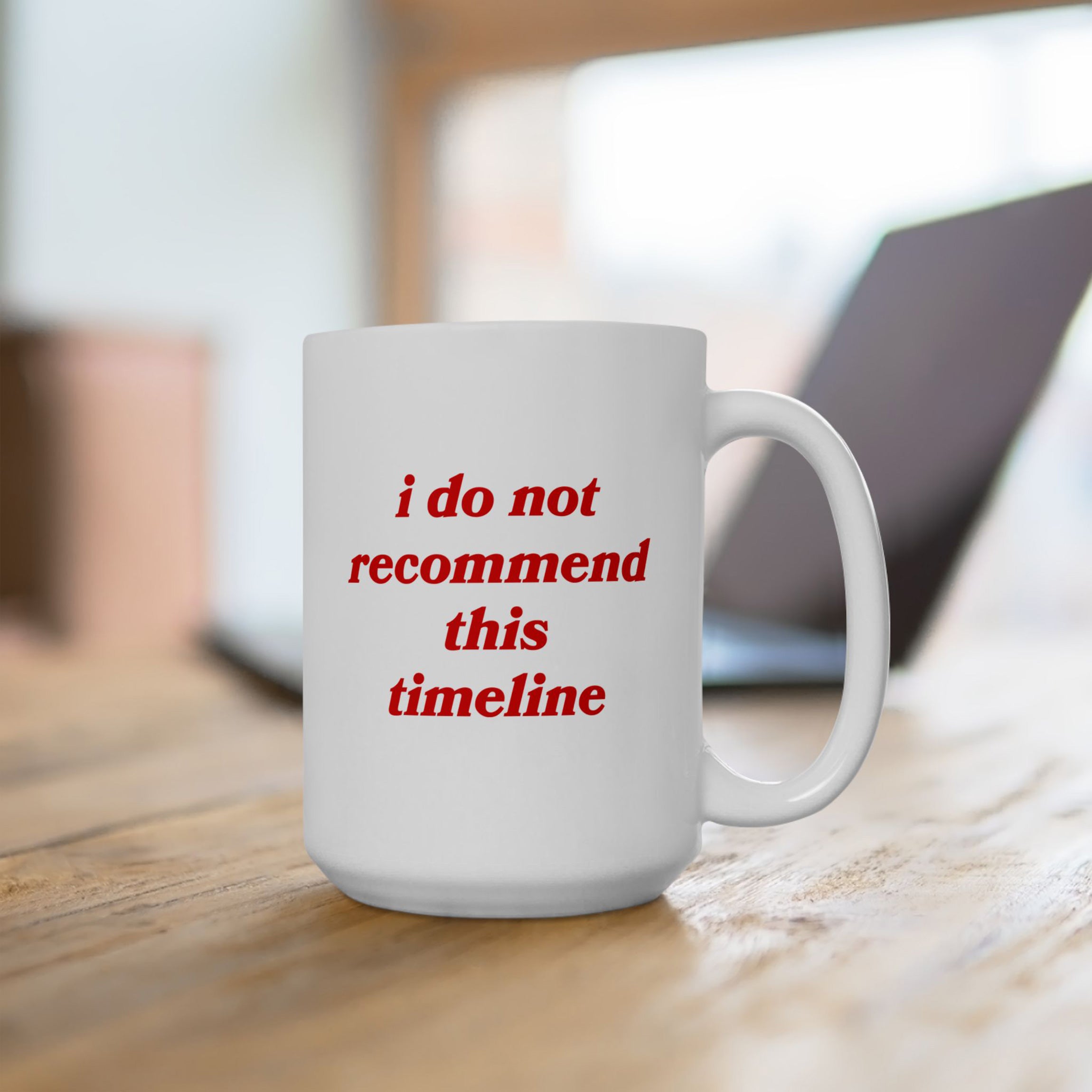 Timeline Mug on wooden table, featuring red serif text, i do not recommend this timeline. Laptop softly blurred in background, perfect for expressing witty discontent and supporting mental health awareness.