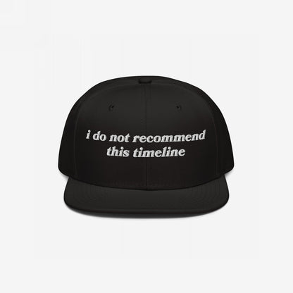 Black Timeline Hat with white embroidery reading, “I do not recommend this timeline.” Ideal for expressing political fatigue and sarcasm. Soft snap-back design against a plain backdrop. Ships from the E.U.