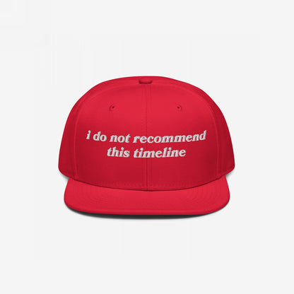 Red flat-bill snapback cap with white embroidered text: “I Do Not Recommend This Timeline.” This Timeline Hat conveys political sarcasm, perfect for expressing discontent with style.