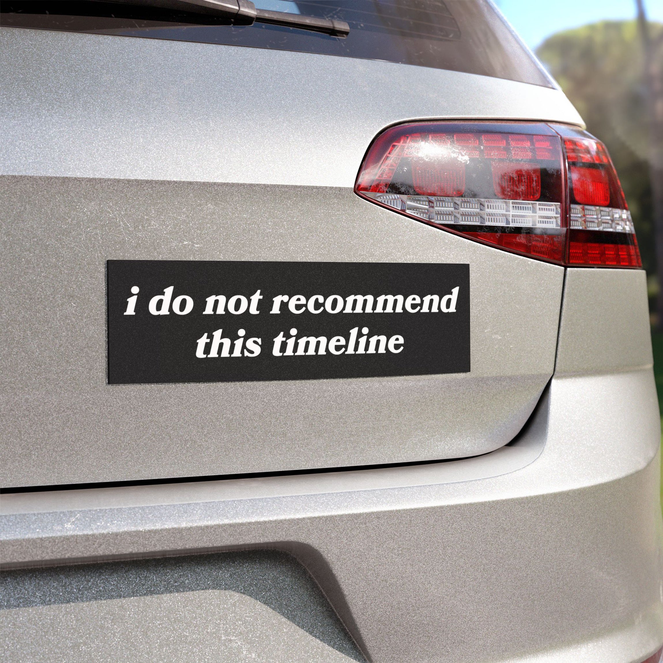 The back of a silver hatchback displays the Timeline Sticker & Magnet with the phrase, “i do not recommend this timeline,” on a black bumper sticker, highlighting existential humor.