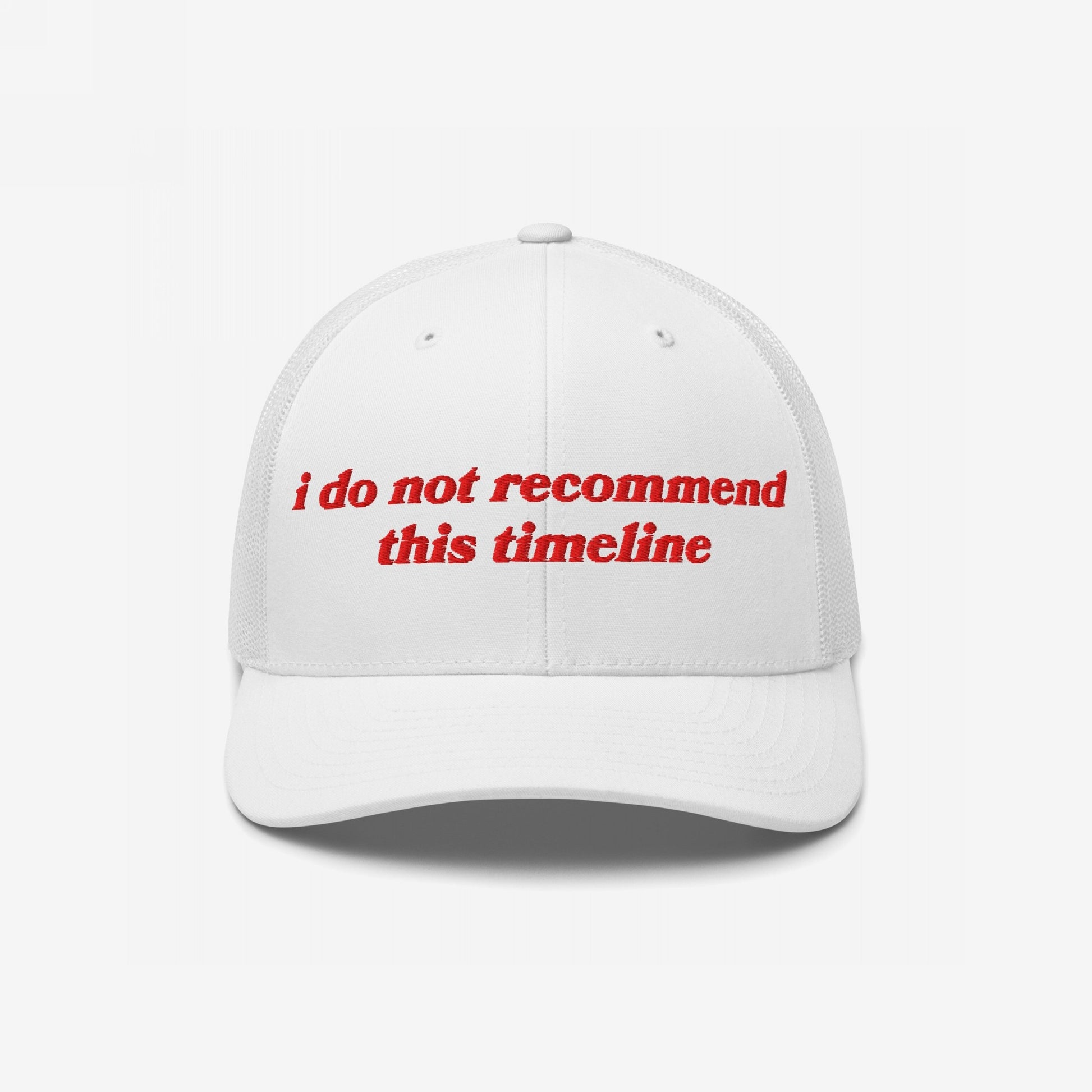 White trucker-style baseball cap with red embroidery reading “I Do Not Recommend This Timeline,” signaling political discontent. Reflects sarcastic, protest-driven ethos, embodying a soft fit for hard truths.