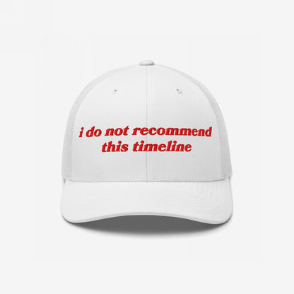 White trucker-style baseball cap with red embroidery reading “I Do Not Recommend This Timeline,” signaling political discontent. Reflects sarcastic, protest-driven ethos, embodying a soft fit for hard truths.