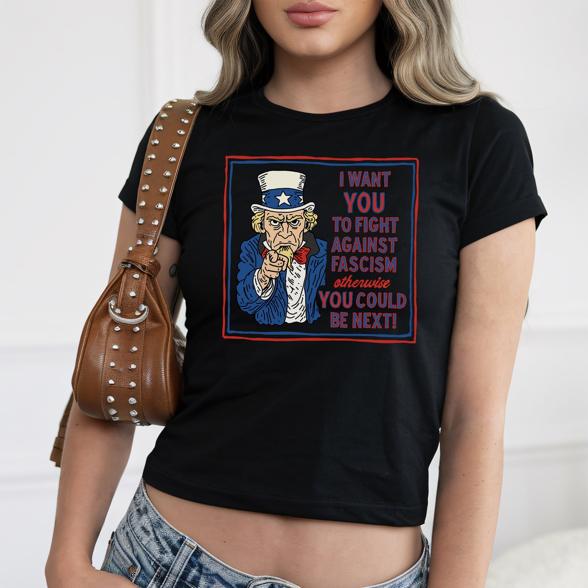 Person wearing Anti-Fascist Uncle Sam Women's Baby Tee with stylized Uncle Sam graphic and bold anti-fascism slogan, paired with studded brown shoulder bag and low-rise blue jeans.