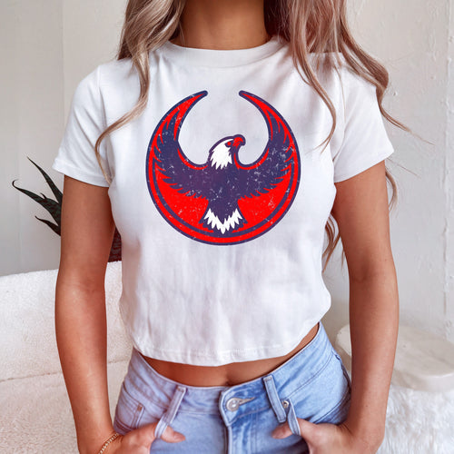 USA Rebel Alliance Women's Baby Tee features a cropped, white T-shirt with a stylized eagle emblem, paired with light-blue high-waisted jeans, embodying a bold stance on immigrant rights and equality.