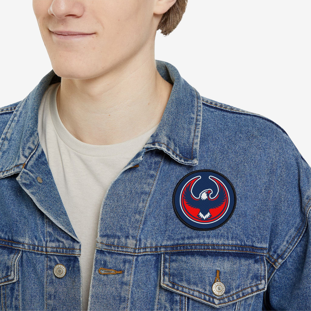 Person wearing a light-wash denim jacket displays a USA Rebel Alliance Patch on the left chest, featuring a stylized eagle, symbolizing anti-fascist and immigrant rights support, over a white T-shirt.