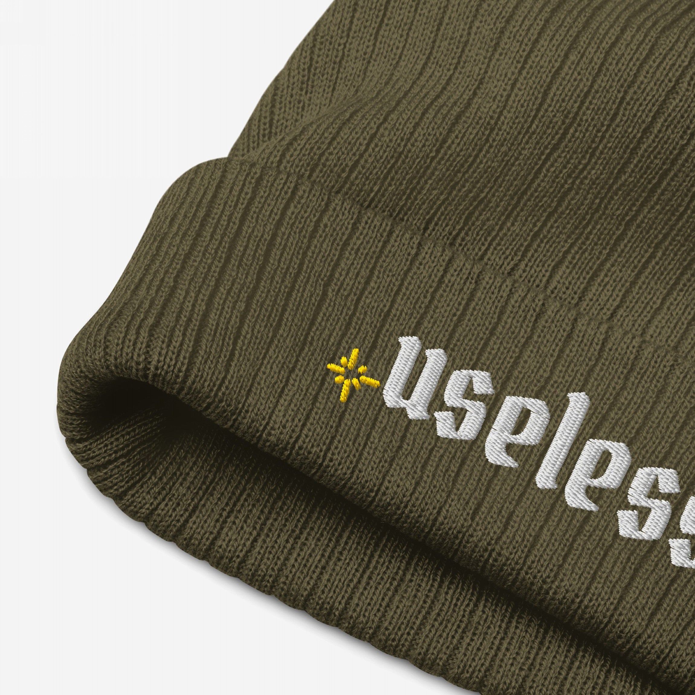 Useless Hat: Close-up of an olive-green rib knit beanie featuring “Useless” in white embroidery on the cuff, with a yellow asterisk logo. A stylish nod to embracing rest and mental well-being.