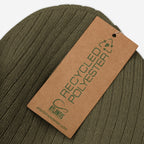 Close-up of the Useless Hat, an olive-green rib-knit beanie with a brown tag marked “RECYCLED POLYESTER” and “ATLANTIS,” embodying a vibe of rest and unapologetic chill.